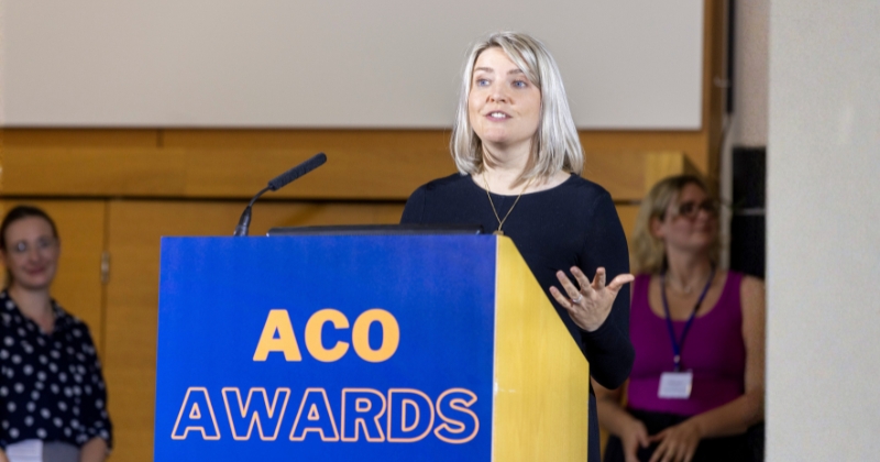 Aileen Edmunds is a woman with blonde hair, dressed in a black top, speaks at a podium during the ACO Awards. The podium is blue and yellow, displaying the words "ACO Awards" in bold orange lettering. She gestures with one hand while addressing the audience, with a microphone positioned on the podium. Aileen is the CEO of Longleigh Foundation.
