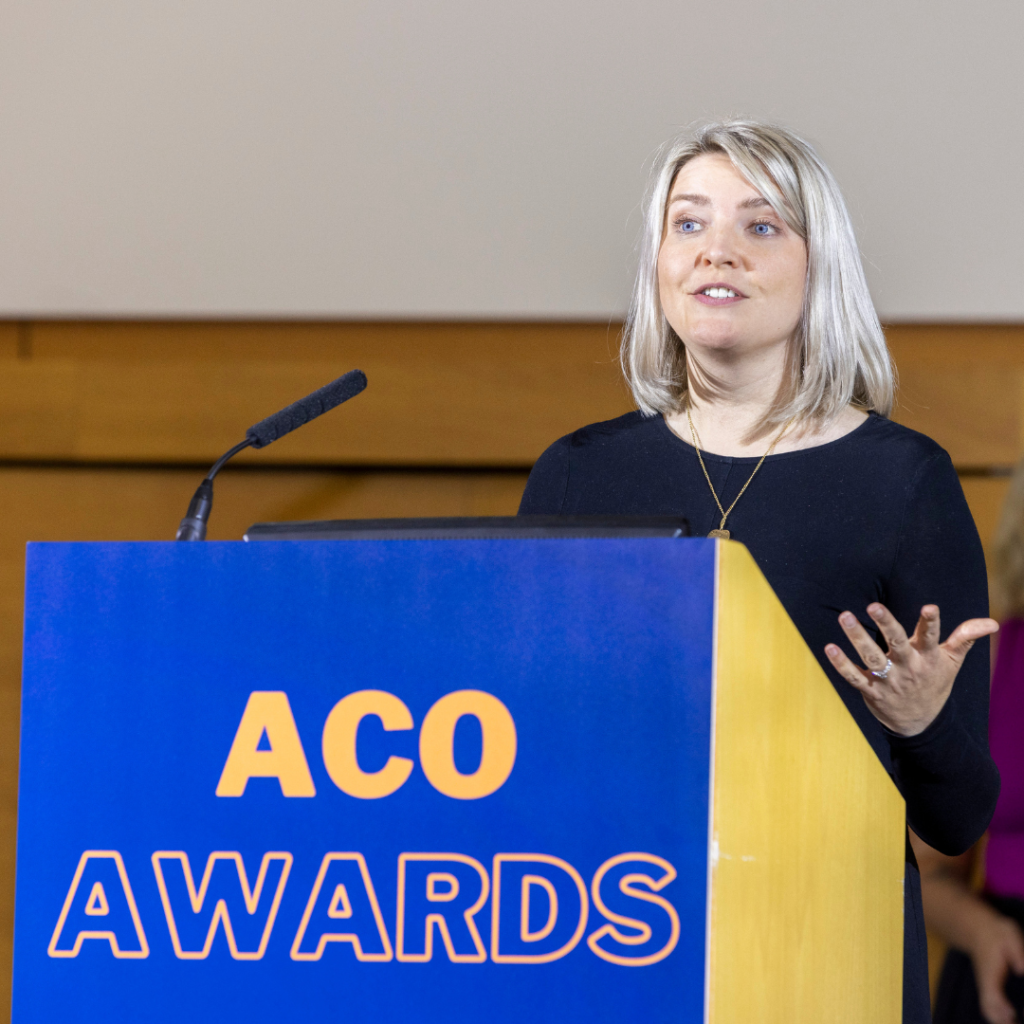 Aileen Edmunds is a woman with blonde hair, dressed in a black top, speaks at a podium during the ACO Awards. The podium is blue and yellow, displaying the words "ACO Awards" in bold orange lettering. She gestures with one hand while addressing the audience, with a microphone positioned on the podium. Aileen is the CEO of Longleigh Foundation.