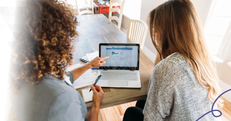 Two people sit at a table looking at a laptop screen with financial data, as one person points at the screen.