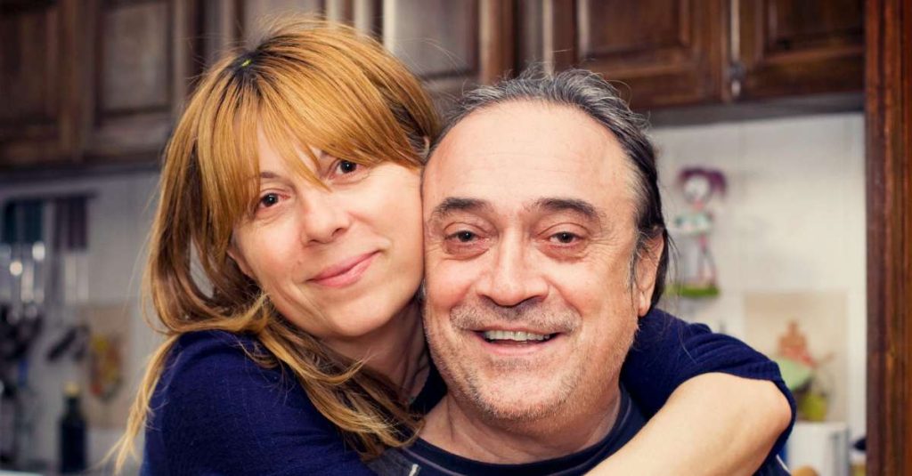 A woman with strawberry blonde hair warmly embraces an older man with graying hair and a cheerful smile. Both are wearing dark blue tops and appear happy and close, standing in a cozy kitchen.