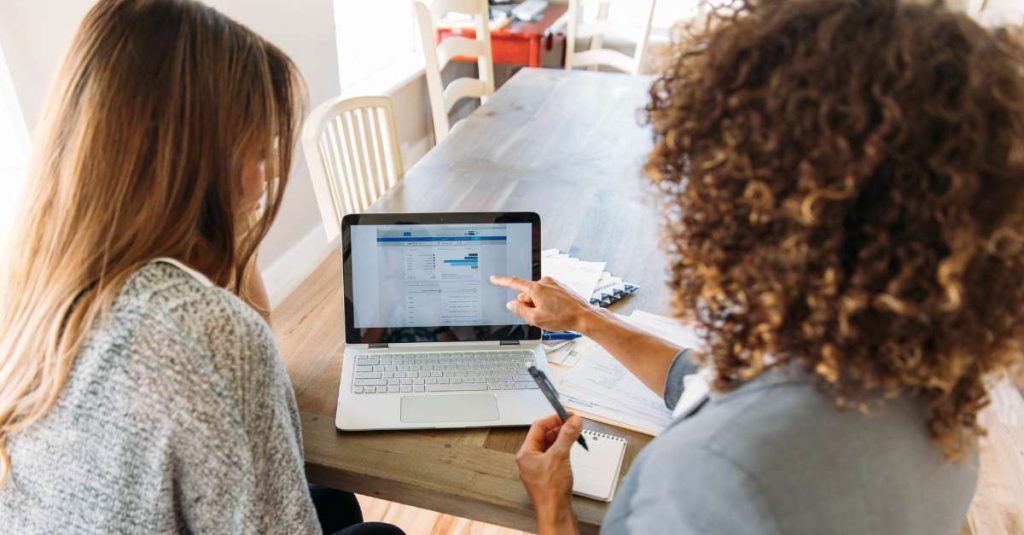 Two women sitting at a wooden table reviewing financial data on a laptop, with one woman pointing at a chart on the screen; documents and a pen are also visible on the table.