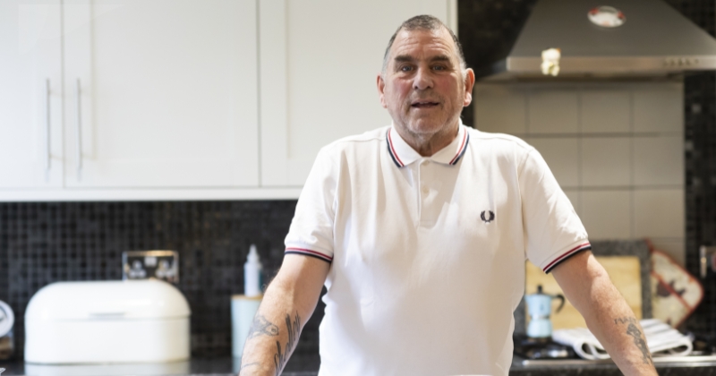 A man stands at a kitchen counter, facing the camera, dressed in a white collared shirt with a small logo. Behind him is a tidy kitchen setting.