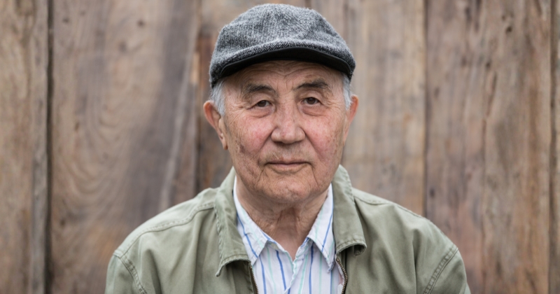 Close-up portrait of an older person wearing a grey flat cap, a light green jacket, and a striped shirt, standing in front of a vertical wooden plank background. The image is centred on the face and upper body, with soft natural lighting and a shallow depth of field that blurs the background.