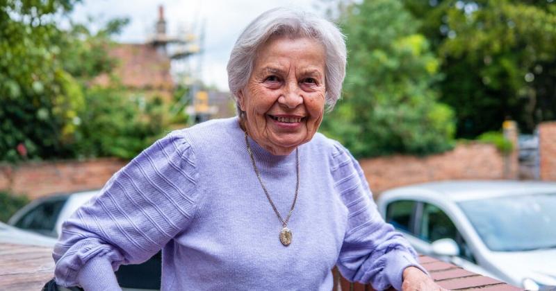 An older lady with short grey hair is standing outdoors, leaning on a surface. The person is wearing a light purple sweater and a gold necklace with a pendant. In the background, there are blurred elements including trees, a brick wall, and part of a car.