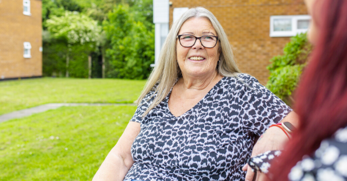 A photo of a lady who has long, shoulder length blonde hair, wearing glasses. She is smiling at a lady who is just off the image and blurred out.
