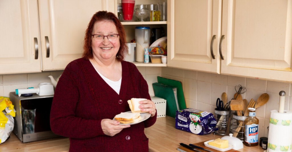 A woman is standing in a kitchen holding a plate with a sandwich and toast, with various kitchen items and food on the countertop behind her.