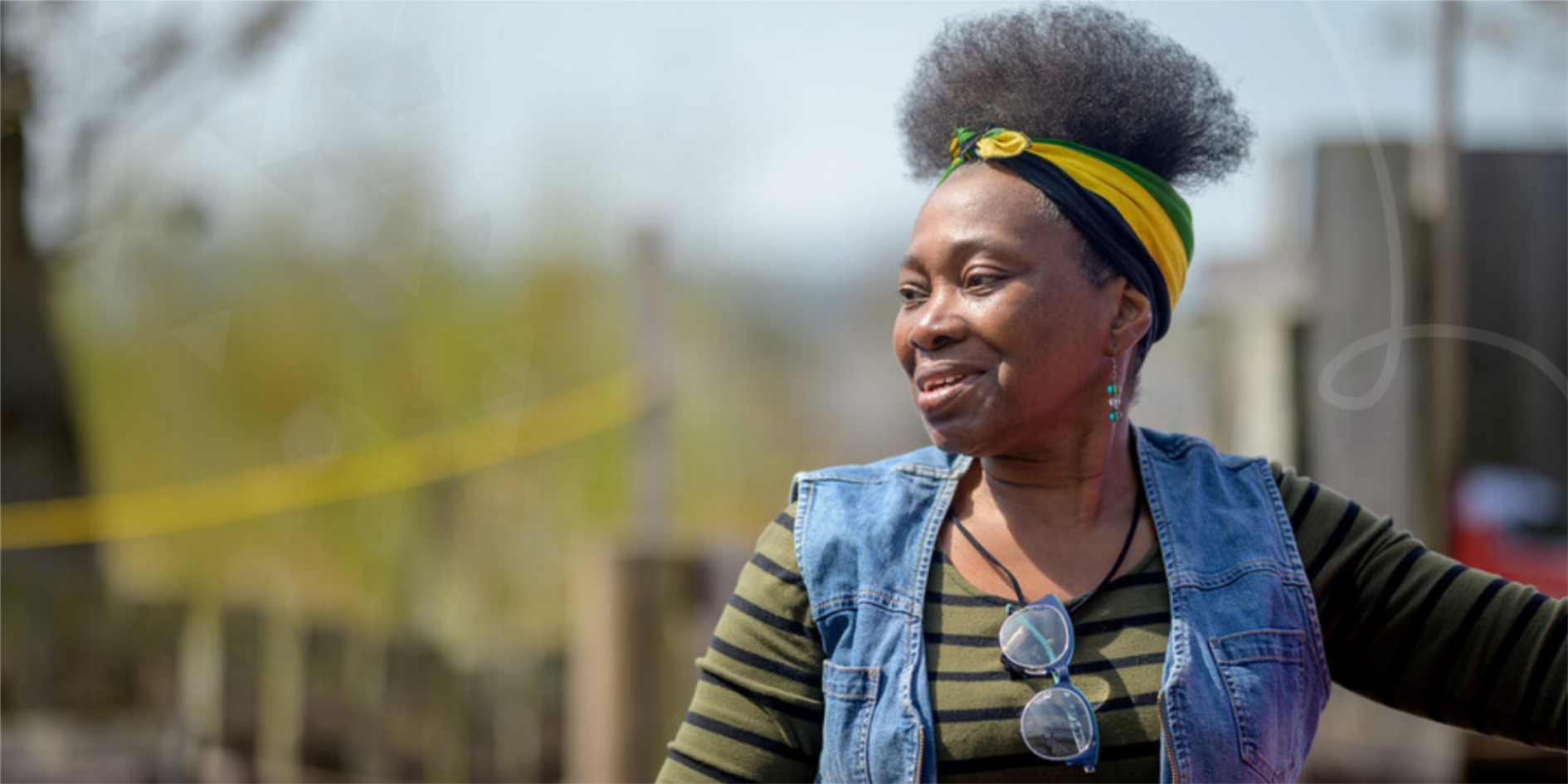 Person standing outdoors wearing a striped top, denim vest, and head wrap, with garden fencing and greenery blurred in the background.