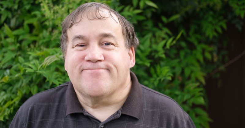 Person wearing a dark polo shirt standing outdoors in front of green foliage.