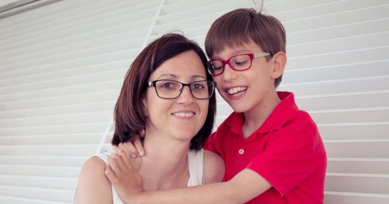 A child held close by his mother in front of white window blinds, with the child’s arms around his mother, symbolising a family gaining the freedom and support needed for safe travel.