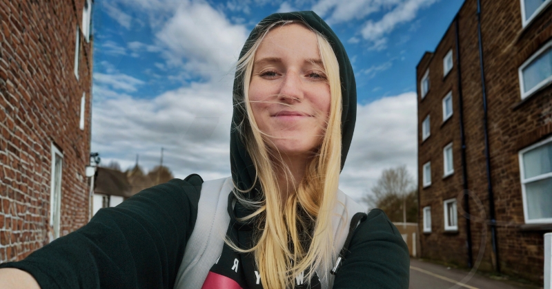A person wearing a dark hooded sweatshirt and light-colored backpack straps takes a close-up selfie outdoors on a residential street. The person’s arm is extended toward the camera, with long light-colored hair blowing across the face. Brick apartment buildings line both sides of the street, and a partly cloudy blue sky fills the background. The image is brightly lit with natural daylight.
