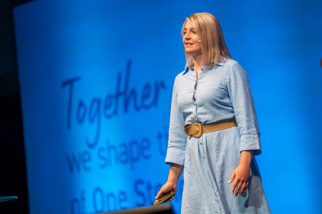 Aileen is speaking on a stage, wearing a light blue dress and holding a presentation remote, with a blue presentation screen in the background.