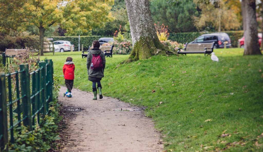 A person wearing a dark coat and backpack walks along a park path beside a young child riding a scooter. The path is bordered by grass, trees, benches, and parked cars in the background on an autumn day.